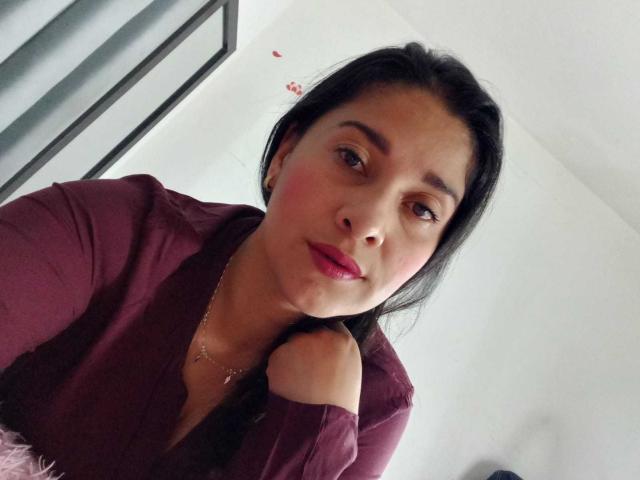 AlanaSteel, com cabelo escuro, batom rosa e olhos castanhos, vestindo uma camisa vinho, segurando o cabelo perto do pescoço, olhando para a câmera com um leve sorriso..