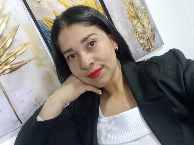 AlanaSteel with red lipstick, black blazer, white ribbed shirt, and pearl earrings, resting her chin on her hand, smiling subtly. Her brown eyes and straight black hair are visible.