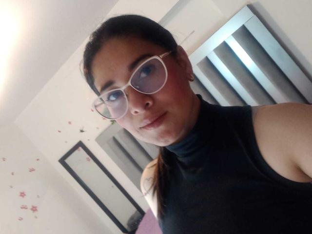 AlanaSteel with glasses, black turtleneck, and ponytail, showing her face and bare shoulders, has a subtle smile and slight blush on her cheeks.