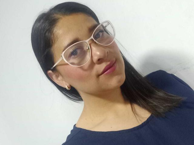 AlanaSteel, usando óculos, com batom rosa e cabelo preto, vestindo uma camisa azul escura, olhando ligeiramente para o lado, sorrindo sutilmente..