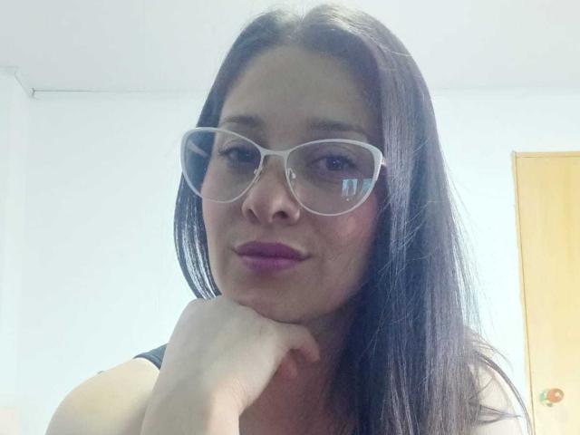 AlanaSteel with long black hair, wearing clear glasses, purple lipstick, and black tank top, resting her chin on her hand, smiling slightly. Her tits are partially visible.