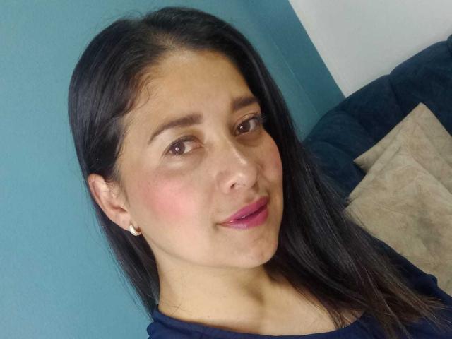 AlanaSteel has long black hair, brown eyes, pink blush, and pink lipstick. She's wearing a blue top and small pearl earrings. Her ass is not visible.