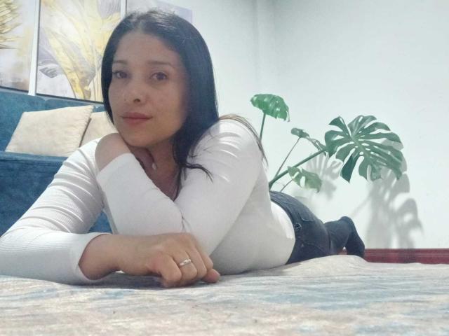 AlanaSteel with black hair, lying on a carpet, wearing a white off-shoulder top, black pants, and a ring, looking at the camera, with a green plant in the background.