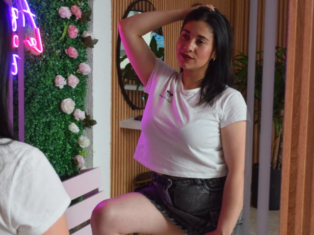 AlanaSteel, with black hair and medium tits, sits in a white t-shirt and black denim skirt, hand in hair, looking to the side with a slight smile.