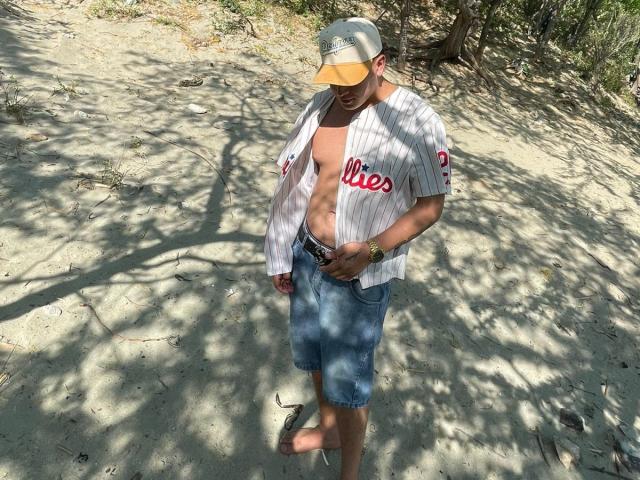 A photograph of ZairaAndres standing on a sandy path. ZairaAndres wears a white pinstriped baseball shirt with "Chis" in red, open to reveal a bare, slightly muscular chest. They also wear blue tie-dye shorts, a black belt, and a yellow baseball cap. ZairaAndres looks down, with one foot raised. Shadows of trees cast on the sandy path. A gold watch is visible on ZairaAndres' wrist. The image is bright and sunny.