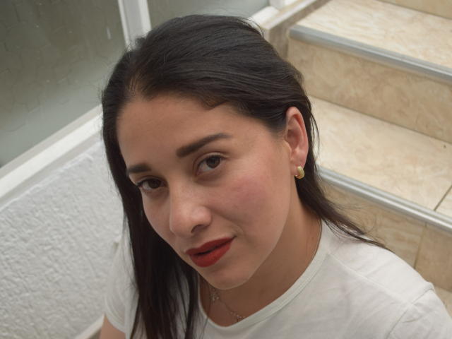 AlanaSteel has a cute face with red lips, dark straight hair, and small earrings. She's wearing a white shirt, standing next to a marble staircase.