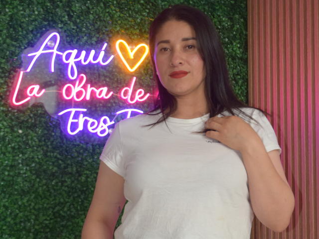 AlanaSteel stands with dark brown hair, white t-shirt, and hand near chest. Neon heart and text on green wall. She's smiling, has red lipstick, and a cute ass.