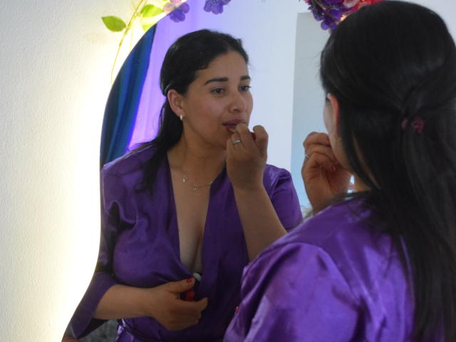 AlanaSteel, con cabello oscuro y vestida con un vestido púrpura que deja ver sus senos, está de pie frente a un espejo, mordiéndose el dedo y mirando su reflejo. Su mano está sobre el vestido..