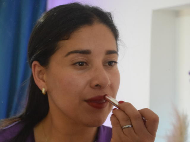 AlanaSteel with dark hair, applying red lipstick with a finger, showing a slight smile. She's wearing a purple shirt and a yellow earring. Her ringed finger is visible.