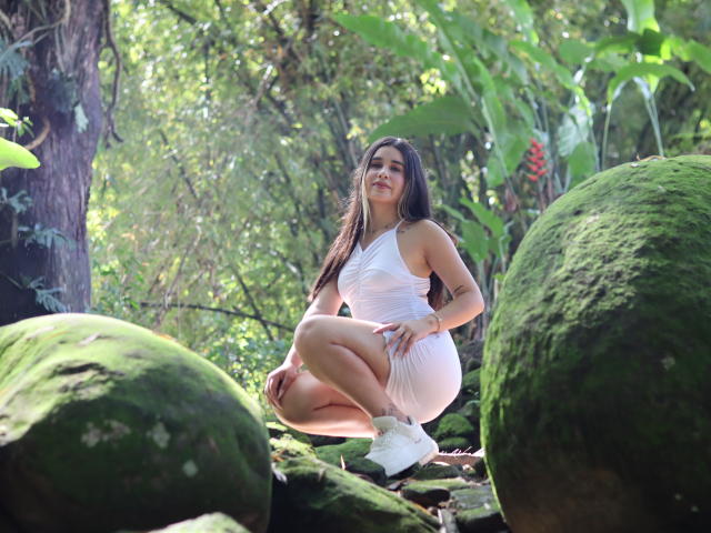 KatieDesirX squatting in a forest, wearing a white one-shoulder dress, showing her tits, with long brown hair, white socks, and green moss-covered rocks in the foreground.