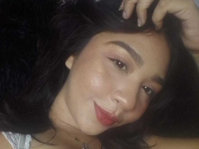 NanaMilan's face with dark hair, brown eyes, and red lipstick, lying down, hand in hair, slight smile, wearing a white top, soft skin, subtle makeup, relaxed vibe.
