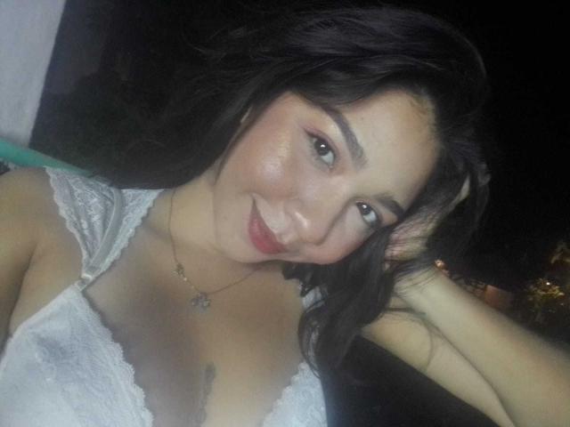 NanaMilan with dark hair, smiling, wearing a white lace top, exposing her small tits, hand in hair, necklace, soft makeup, light skin, slightly flushed, night setting.