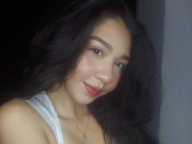 NanaMilan with long black hair, light brown skin, red lipstick, subtle makeup, white lace top, and a small necklace, smiling softly at the camera.