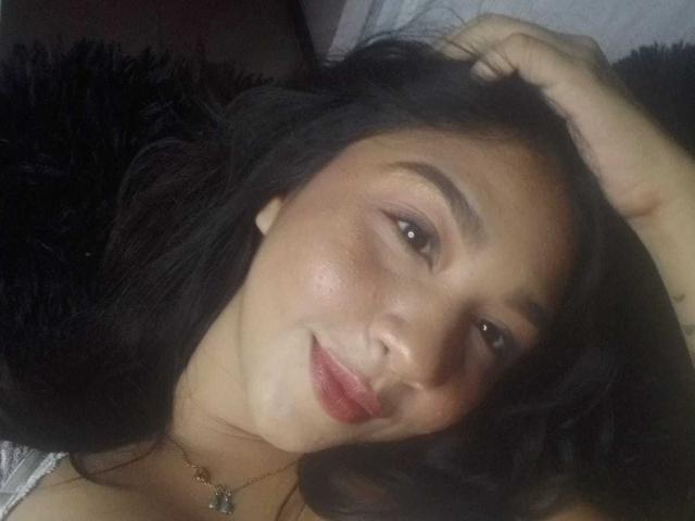 NanaMilan with dark hair, brown eyes, and red lipstick, lying down, hand in hair, smiling slightly, wearing a necklace, soft skin, black fuzzy pillow in background.