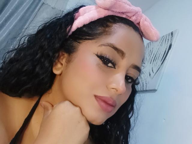 MariaCatalina with curly black hair, pink headband, and long eyelashes, resting her chin on her hand, showing her pouty pink lips, and her small, round nose.