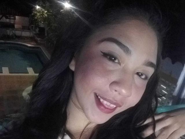 NanaMilan with dark hair, full lips, and winged eyeliner, smiling at the camera, showing her teeth, under bright lights, in a night setting. Her ass is not visible.