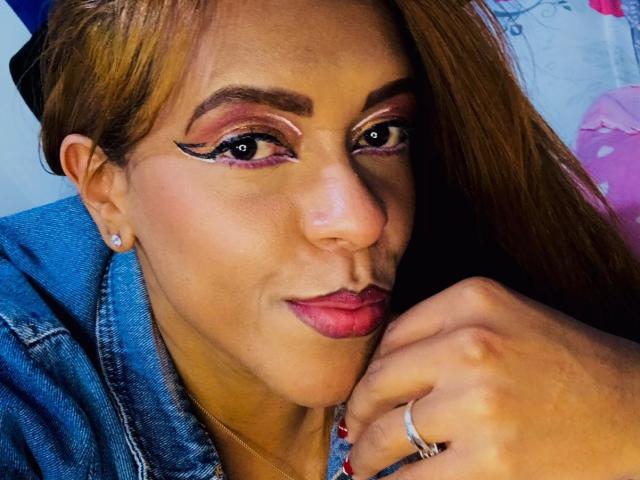 ShamanthaFire has brown hair, bold pink eyeliner, and purple lipstick. She's wearing a denim jacket, diamond earrings, and a silver ring on her finger. She's smiling softly.
