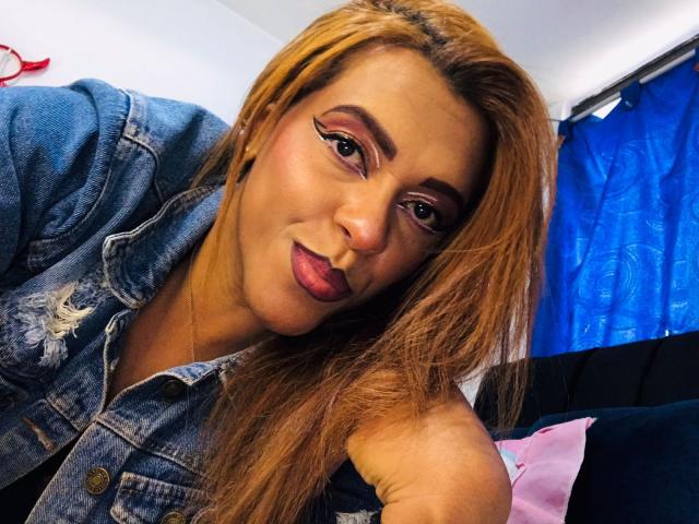 ShamanthaFire with pouty lips, wearing a denim jacket, has long blonde hair, bold eyeliner, and pink lipstick, leaning forward in a selfie. Her blue curtain is in the background.