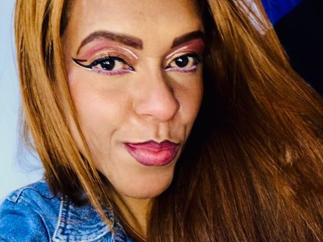 ShamanthaFire with long brown hair, pink eyeliner, and pink lipstick, wearing a blue denim shirt, has a slight smirk and looks directly at the camera.