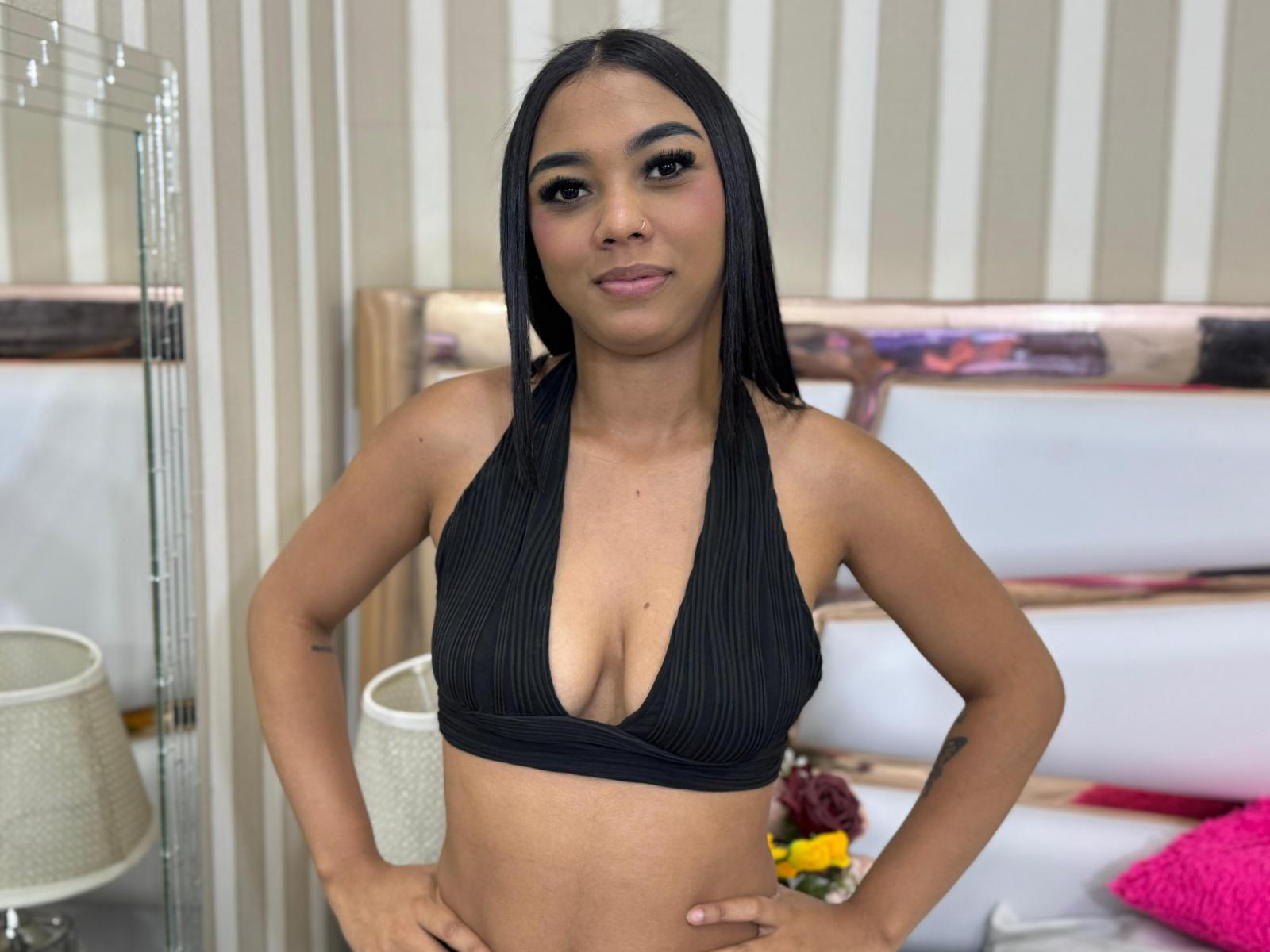 TamaraRochers with straight black hair, brown eyes, and tan skin, wearing a black halter top that reveals her small tits, standing with hands on hips, smiling.