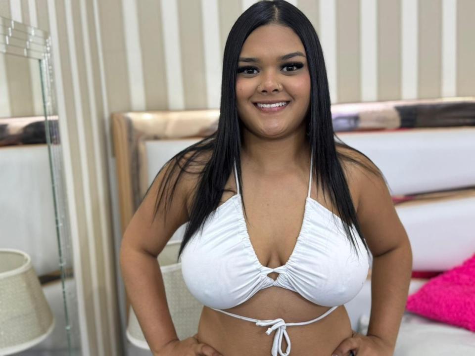 EmilyRochet is a young woman with long black hair, smiling at the camera. She wears a white, halter-style bikini top that accentuates her large breasts. Her hands are on her hips. The background includes a striped wall, a mirror, and a bed with a white headboard and pink pillow. The image is brightly lit, focusing on EmilyRochet's cheerful expression and her upper body. Her skin is a warm brown tone.