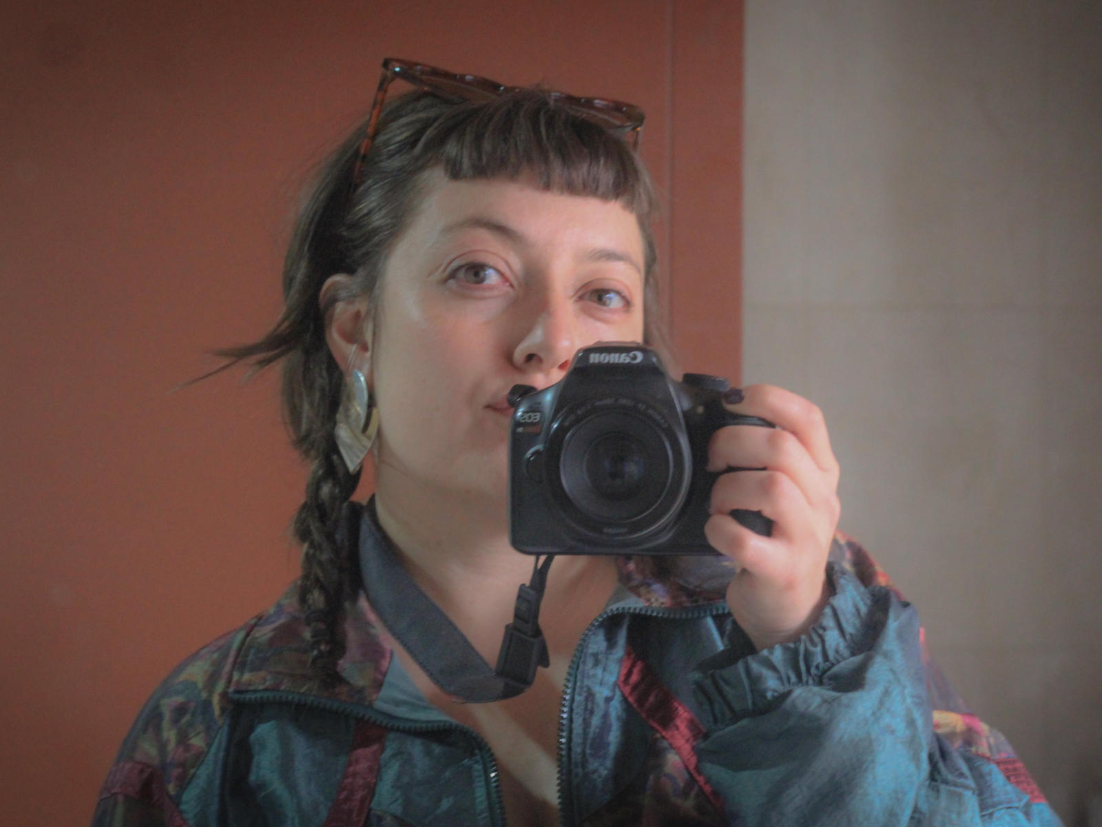 SashaGrano takes a selfie with a Canon camera. She has a short, messy brown hairstyle with braids, and wears a patterned jacket. Her face is centered, slightly turned to the right, with a subtle smile. Sunglasses rest on her head. The camera is held close to her face, covering her nose. The background is a simple, two-toned wall. The image is slightly out of focus, emphasizing the casual, spontaneous nature of the selfie.