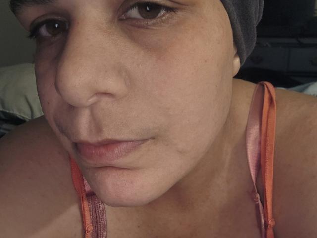 Close-up of CockTease's face. Light brown skin, slight blemishes, small nose, and brown eyes. Grey headwrap, brown eyes with subtle dark circles. Wearing an orange tank top with thin straps. Soft, natural light, slightly shadowed. Gentle, neutral expression. Background is blurred, dark. Image focuses on facial details and tank top. Simple, realistic composition.