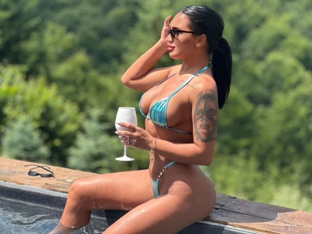 SarahAdams with wet skin, blue bikini, large boobs, tattooed arm, holding a wine glass, sitting by a hot tub, dark hair in a ponytail, wearing sunglasses, sunny day.