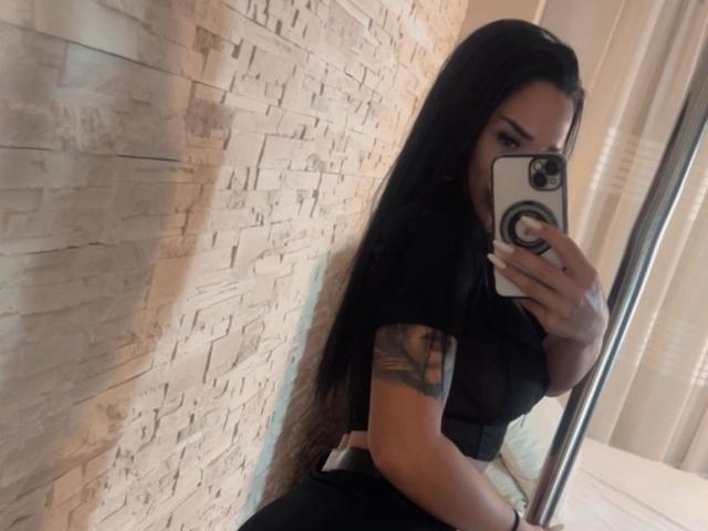 SarahAdams with long black hair, black sheer top, tattoo on right arm, holding phone, mirror selfie, showing her tits, standing against textured wall, holding phone with ring.