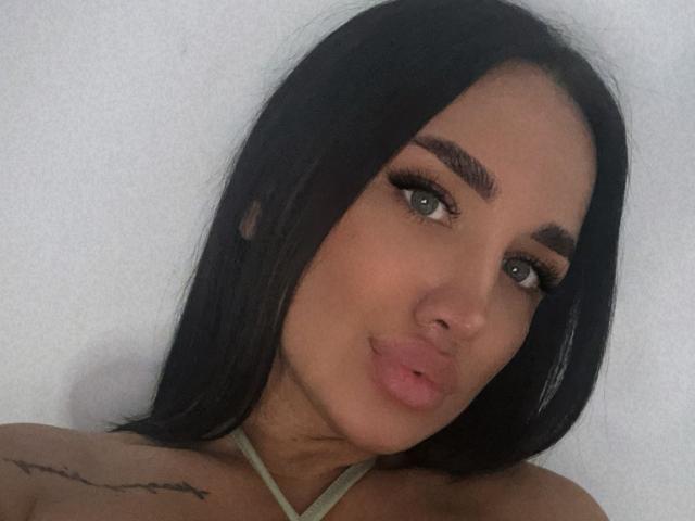 SarahAdams with long black hair, full eyebrows, and pouty lips, has a small tattoo on her right shoulder and is wearing a green halter top. Her tits are visible.