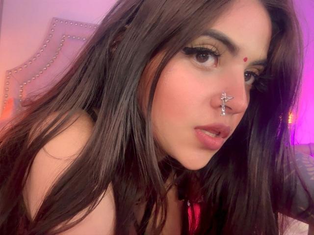 CoriPaige has long brown hair, green eyes, and wears a nose ring with a star charm. Her red dot is on her forehead, and she has long, thick eyelashes.