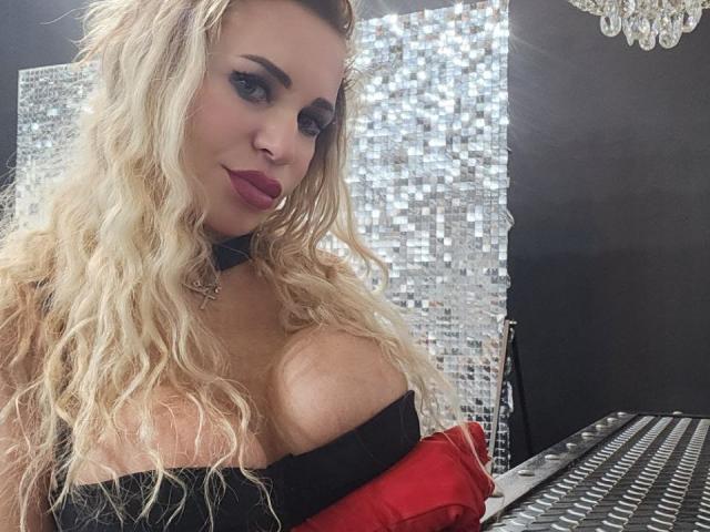 PamellaaOh has blonde wavy hair, blue eyeshadow, red lipstick, black choker, large boobs in a black and red strapless top, sitting at a reflective table.