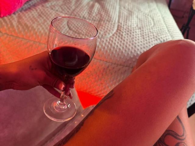 La main d'Amberv tient un verre de vin rouge, révélant des tatouages sur sa cuisse et son genou. Elle est assise sur un lit avec une couverture blanche texturée..