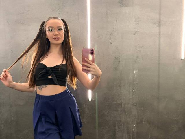 MistyReeds with long brown hair, glasses, black tied-up crop top, and blue pants, holding phone in mirror selfie, showing tattoo on left side, holding hair with right hand.