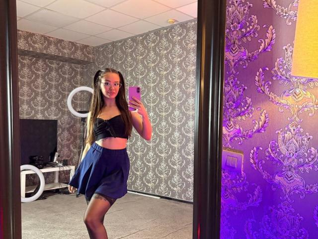 MistyReeds stands in front of a mirror, holding a phone, wearing a black strapless top and blue skirt, with long brown hair, and a cute face, showing her ass.