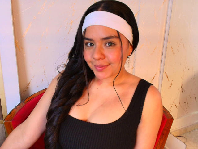 ArielleWilde with long black hair, white headband, black ribbed tank top, smiling, showing small tits, sitting on red chair, light skin, cute, earbuds in.