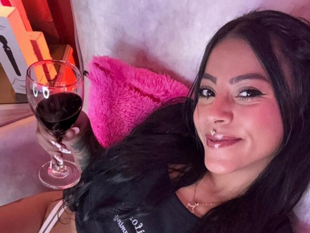 Amberv mit schwarzen Haaren, voller Lippen und einem Nasenring, hält ein Glas Rotwein, trägt ein schwarzes Hemd und hat ein pinkes Kissen hinter sich..