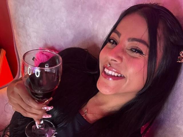 Close-up of Amberv with long black hair, smiling with pink lipstick and a nose ring. She holds a glass of red wine with a pink drink float in it. Her nails are long with white tips. She wears a black top and a thin necklace. The background is a white, textured surface with a red-orange object on the left. The image has a casual, candid feel.