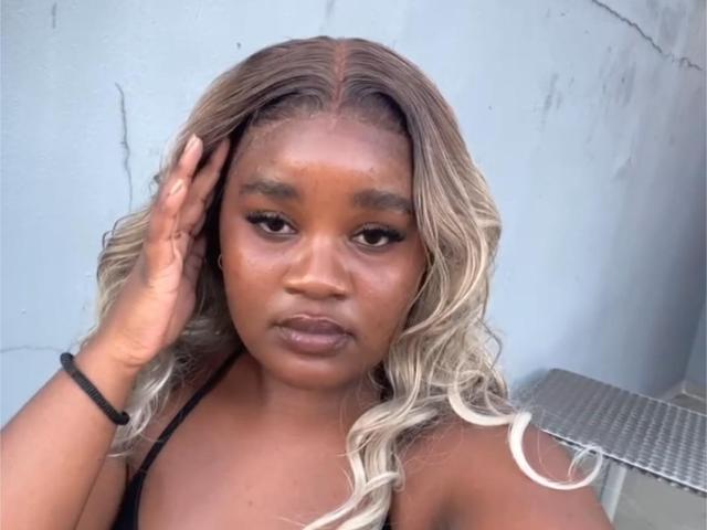 StellaJoanne has dark brown skin and wavy blonde hair with dark roots. She's looking at the camera with a neutral expression, her right hand touching her hair. She's wearing a black strap top. Her makeup includes dark eyeliner and subtle lip color. She has a black bracelet on her right wrist. The image has a casual, selfie-style composition. The background is a plain, light blue wall. The lighting is even, highlighting her face and hair.