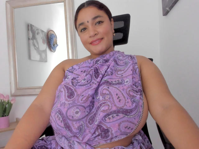 DarsshaHara with big tits in a purple paisley dress, sitting in a black chair, smiling, brown hair in a bun, fair skin, light makeup, and a pink flower vase.