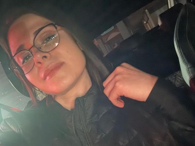 LustEncrypted with glasses, dark hood, and black puffy jacket, slightly flushed face, hand near chin, semi-smiling, brown hair, dim lighting, visible car seat.