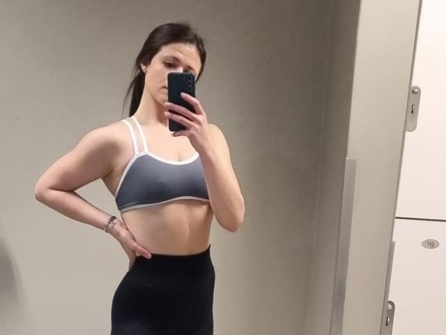 A selfie of LustEncrypted with a mirror. She has long dark brown hair, fair skin, and a toned physique. She wears a gray sports bra with white straps and black high-waist leggings. She holds a black smartphone in her right hand, slightly covering her face. Her left hand is on her hip. The room has a neutral gray wall and white locker on the right. The image is clear and focuses on her upper body.