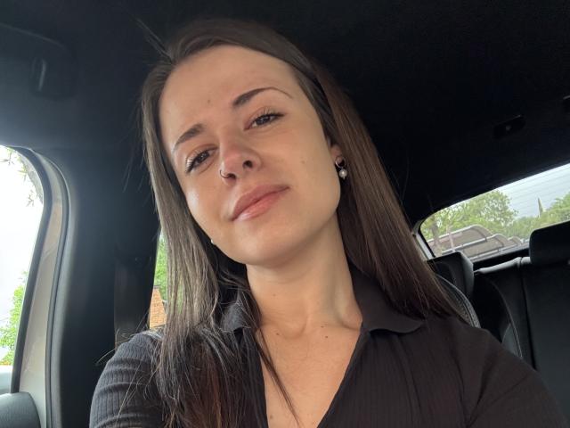 Close-up selfie of LustEncrypted, a young woman with light skin, straight brown hair, and a small nose ring in her left nostril. She has thin eyebrows, subtle eyeliner, and neutral lipstick. She wears a black, ribbed, collared shirt and small, pearl stud earrings. Her expression is neutral, slightly smirking. The car's interior is visible, with a black roof and window on the right showing green foliage and a house roof. Natural light enters from the window. The composition is centered, with a slight upward angle.