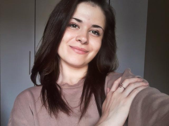 LustEncrypted, a cute chick with dark brown hair, smirks at the camera, wearing a pinkish-brown sweater, hand on shoulder, ring visible, soft natural light, slightly flushed face.