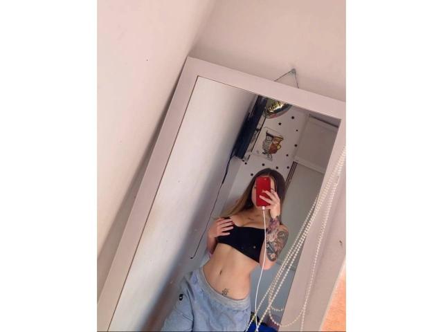 LunaBaster with small tits, wearing a black crop top and loose gray pants, taking a mirror selfie with a red phone, showing a tattoo on her right arm.
