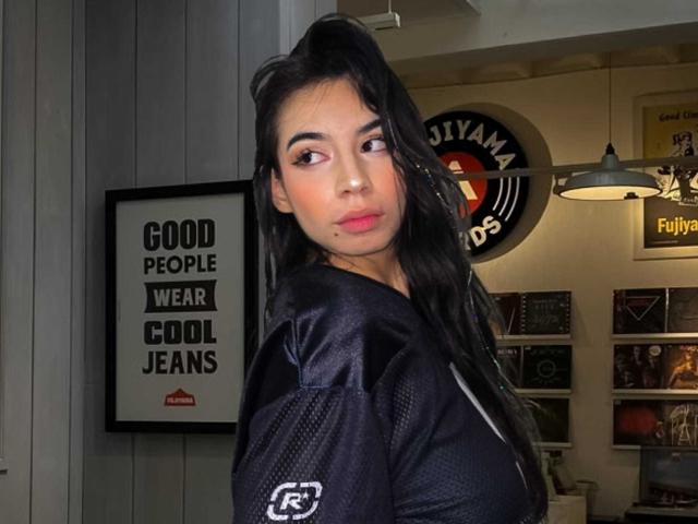 KatherinJohns with long black hair, wearing a black sports jersey, looking over her shoulder, medium tits, slight smile, in a dimly lit room with wall art.