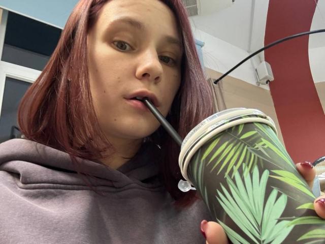 MonaTrio with red hair, brown eyes, and a grey hoodie, sucking on a straw from a green leaf-patterned cup, holding it with red-painted nails.