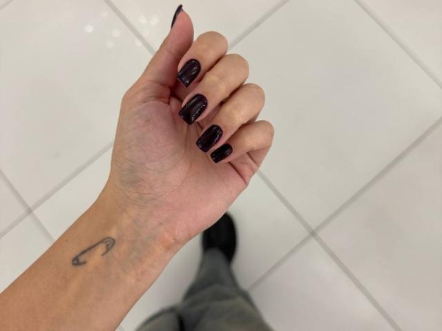 Die Hand von MonaTrio mit schwarzen, glitzernden Nägeln, ein sichtbares Tattoo in Form einer geschwungenen Linie am Handgelenk, ein weiß gefliester Boden und graue Hosen im unteren Teil des Bildes..