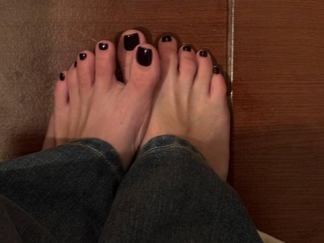 MonaTrio's feet with dark black painted toenails, resting on a wooden floor, wearing blue jeans. Her toes are slightly curled. Her skin is fair.