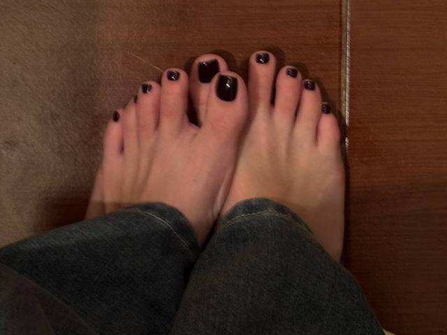 Os pés de MonaTrio, com esmalte preto escuro nos dedos, vestindo jeans azuis, em pé em um piso de madeira. Seus dedos estão ligeiramente curvados, mostrando a textura da madeira..